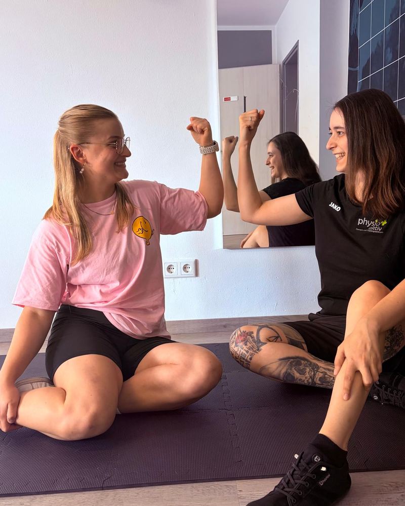 Zwei Therapeutinnen von ErgoPlus sitzen auf einer Trainingsmatte und zeigen sich lachend den angespannten Oberarm