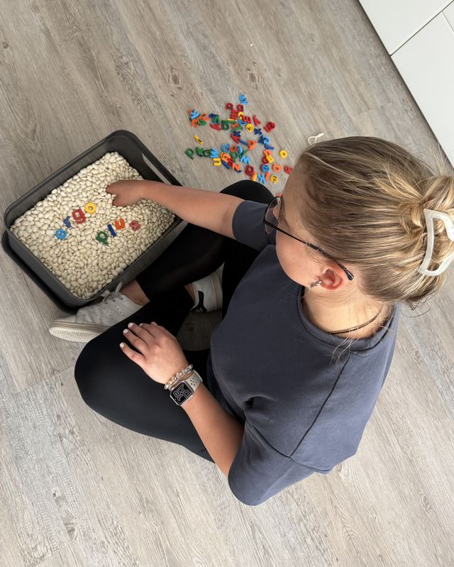 Eine Therapeutin sitzt auf dem Boden neben einer Kiste mit weißen Bohnen, darin liegen bunte Buchstaben, die ergo plus ergeben