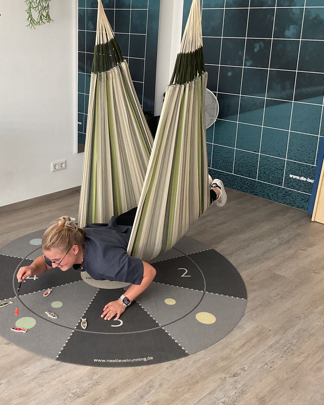 Eine Therapeutin liegt bäuchlings in einer Therapiehängematte und greift nach Spielfiguren auf einer Bodenmatte