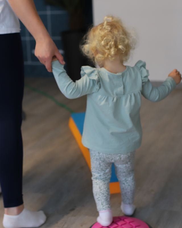 Ein Kleinkind balanciert an der Hand einer Therapeutin über ein weiches Übungsgerät