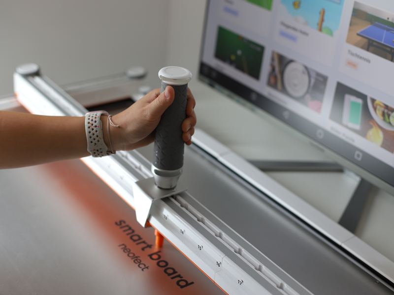 Eine Hand führt den Griff des Neofect Smart Board, dahinter zeigt ein Bildschirm Übungsspiele
