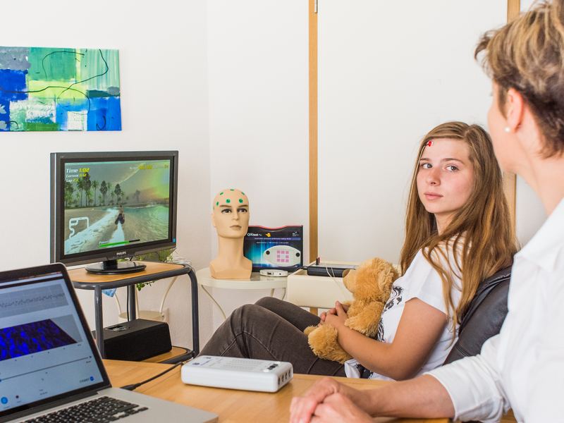 Eine Jugendliche mit Elektroden am Kopf sitzt beim Neurofeedback vor einem Bildschirm mit Spiel, daneben eine Therapeutin und ein Laptop mit Auswertung
