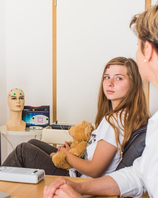 Eine Jugendliche mit Elektroden am Kopf trainiert beim Neurofeedback, neben ihr ein Elektrodenmodell und eine Therapeutin