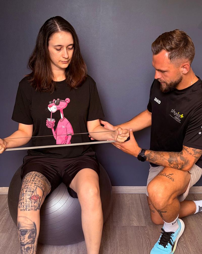 Ein Physiotherapeut von PhysioPlus kniet neben einer Patientin auf dem Pezziball und korrigiert ihre Übung mit dem Theraband
