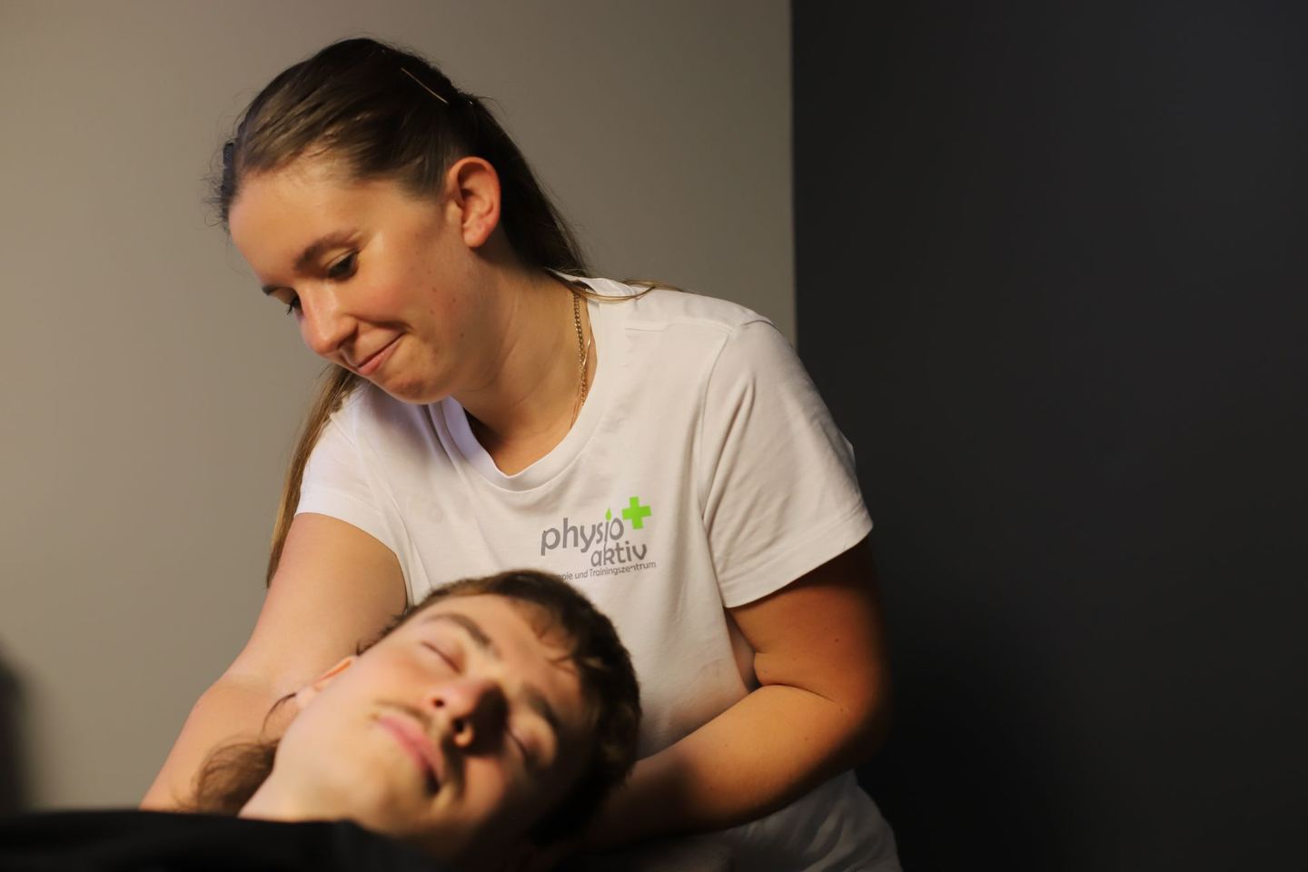 Eine Physiotherapeutin behandelt lächelnd einen liegenden Patienten an Kopf und Nacken